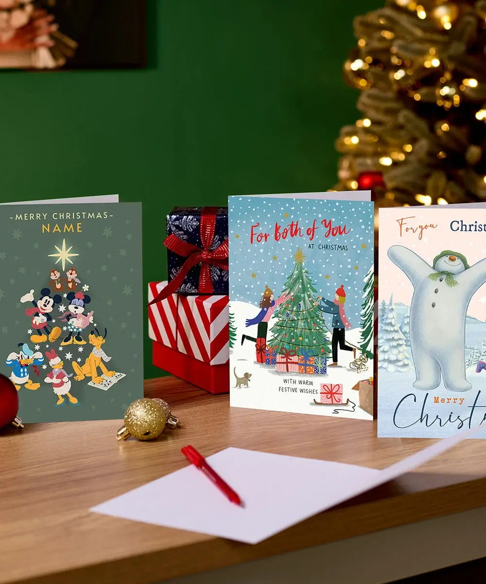 Selection of Christmas cards, including Disney, festive and personalised designs, displayed on a wooden table with wrapped presents and a decorated Christmas tree in the background. Selection of Christmas cards, including Disney, festive and personalised designs, displayed on a wooden table with wrapped presents and a decorated Christmas tree in the background.